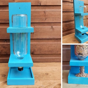 May include: A blue wooden bird feeder with a clear glass bottle for holding birdseed. The feeder has two shelves and a tray at the bottom for catching spilled seed.