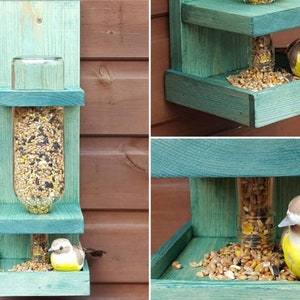 May include: A teal blue wooden bird feeder with two platforms. The feeder has a clear glass bottle in the center with birdseed inside. A small yellow bird figurine is perched on the bottom platform.