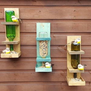 May include: Three wooden bird feeders with glass bottles attached. The feeders are mounted on a wooden wall. The bottles are filled with birdseed and there are small toy birds perched on the feeders.