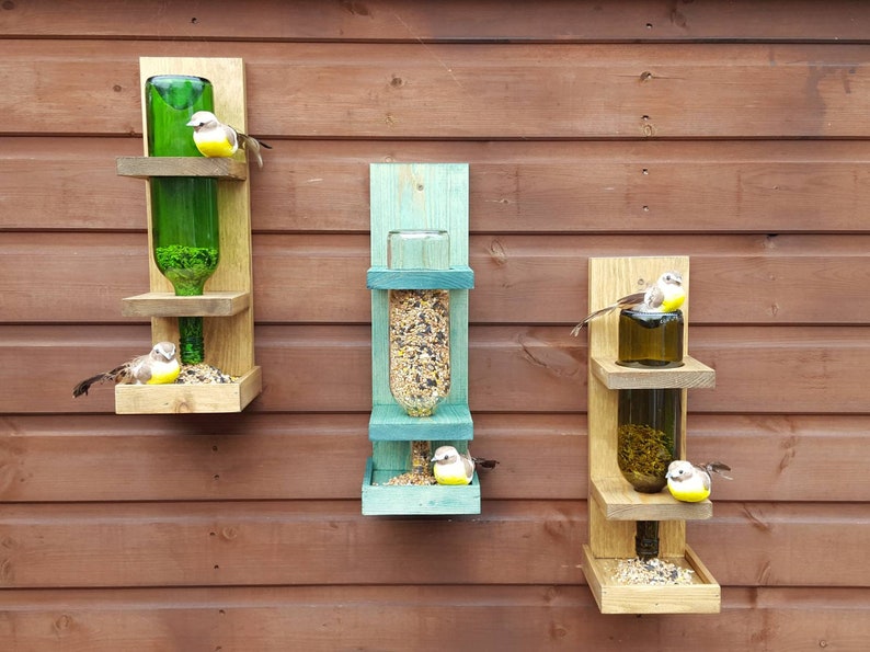 May include: Three wooden bird feeders with glass bottles attached. The feeders are mounted on a wooden wall. The bottles are filled with birdseed and there are small toy birds perched on the feeders.