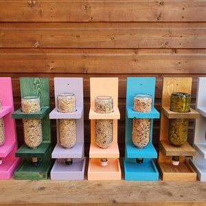May include: Eight bird feeders made from repurposed glass bottles. Each feeder is painted a different color: pink, green, purple, orange, blue, brown, white, and gray. The feeders are mounted on a wooden wall and have a platform below for birds to perch.