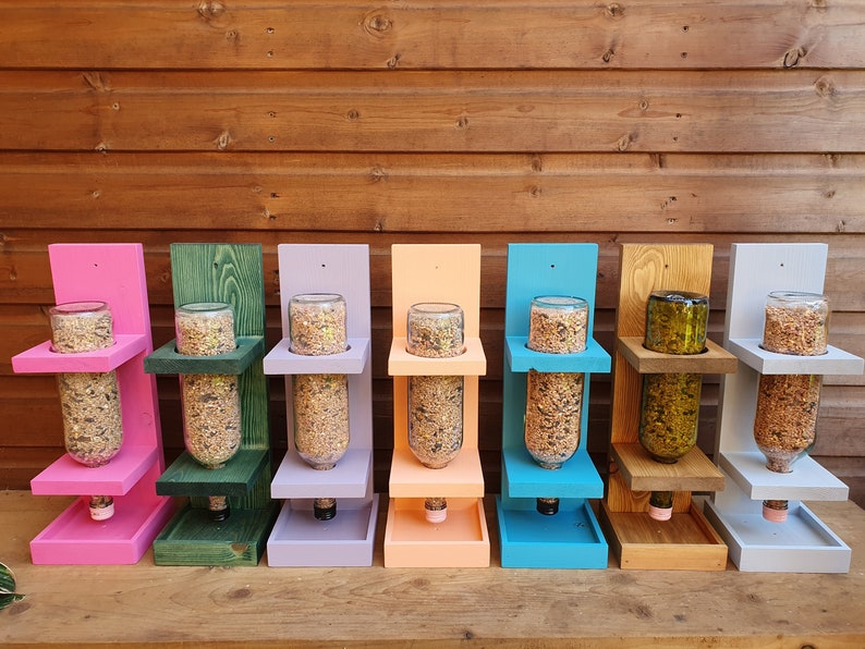 May include: Eight bird feeders made from repurposed glass bottles. Each feeder is painted a different color: pink, green, purple, orange, blue, brown, white, and gray. The feeders are mounted on a wooden wall and have a platform below for birds to perch.
