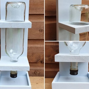May include: A white wooden bird feeder with two shelves and a clear glass bottle. The bottle has a black cap and is attached to the bottom shelf. The feeder is designed to hold birdseed and has a tray at the bottom to catch any spills.