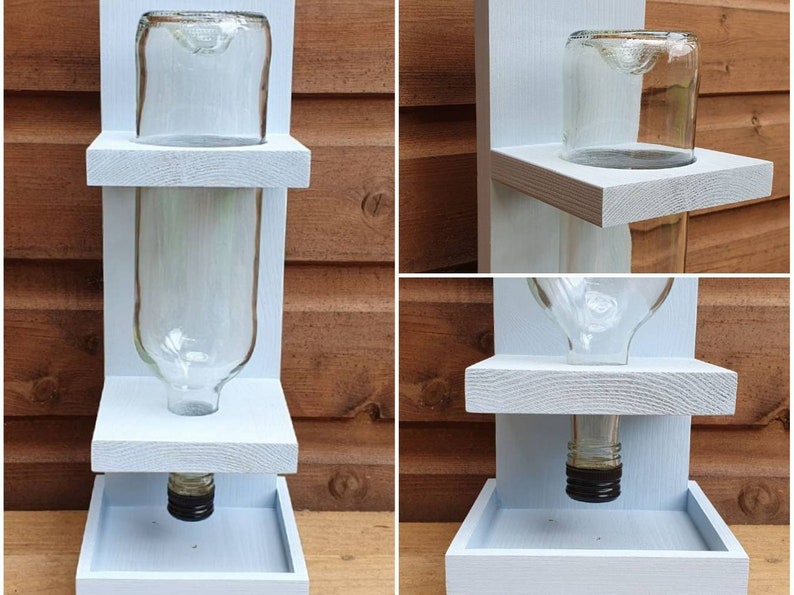 May include: A white wooden bird feeder with two shelves and a clear glass bottle. The bottle has a black cap and is attached to the bottom shelf. The feeder is designed to hold birdseed and has a tray at the bottom to catch any spills.