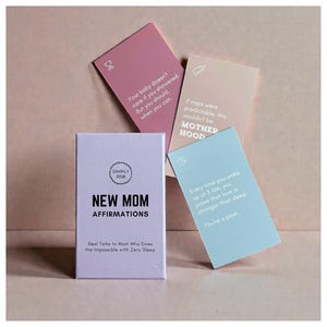 May include: A set of affirmation cards for new mothers, with a lavender box reading "NEW MOM AFFIRMATIONS." The cards are in pastel colors, with phrases like "Your baby doesn't care if you showered" and "Every time you wake up at 3 AM, you prove that love is stronger than sleep."