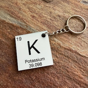 以下が含まれることがあります： カリウムの化学元素記号Kが記された正方形の白いキーホルダー。左上隅に原子番号19、下部に原子量39.098が黒い文字で印刷されており、銀色の金属製丸型二重リングに取り付けられています。