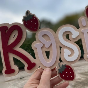 Könnte beinhalten: Ein Holzschild mit dem Namen "Rosie", der in roten, weißen und rosafarbenen Buchstaben geschrieben ist. Die Buchstaben sind in Braun umrandet und haben einen verspielten, handgezeichneten Stil. Auf dem Schild befinden sich drei rote Erdbeeren mit grünen Blättern.