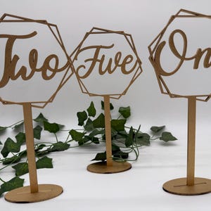 May include: Three wooden table number signs. Each sign has a geometric frame with a number in a cursive font. The numbers are "Two", "Five", and "One". The signs are on a stand with a round base. Green leaves are in the background.