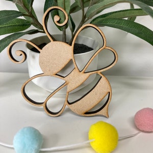 May include: A wooden cutout of a bee with a brown outline. The bee is in a simple, cartoon style with a rounded body and wings. The bee is sitting on a white surface.