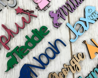 May include: A collection of wooden name plaques with various colors and designs. The names include Teddy, Noah, Millie, Jax, George, and others. Each plaque features a small graphic, such as a rocket, a star, a tent, a unicorn, and a pig.