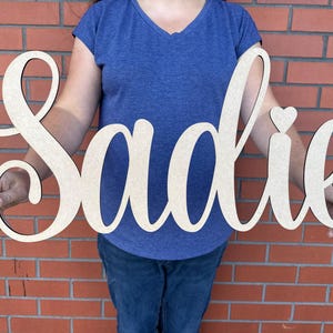 May include: A wooden sign spelling "Sadie" in a cursive font. The sign is made of natural wood with a dark outline and a small heart above the letter "i". The sign is held in front of a brick wall.