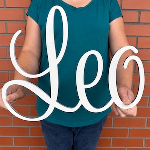 May include: A white wooden sign spelling "Leo" in cursive script. The sign is held by a person against a brick wall. The sign is a decorative item, suitable for home decor.