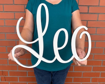 May include: A white wooden sign that spells out the name "Leo" in a cursive font. The sign is held by a person in front of a brick wall. The sign is a decorative item.