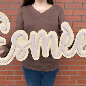 Peut inclure: Panneau en bois avec le nom "Esmée" dans un design superposé. La couche supérieure est rose clair et la couche inférieure est en grain de bois naturel. Le panneau est tenu devant un mur de briques.