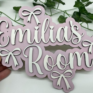May include: A pink wooden sign with the words "Emilia's Room" in white script. The sign is decorated with white bows and hearts. A decorative item for a child's bedroom.