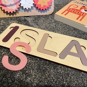 Puede incluir: Un rompecabezas de madera con el nombre "ISLA" recortado. Las letras son de diferentes tonos de marr&oacute;n y rosa. La "S" es una pieza separada y es rosa.