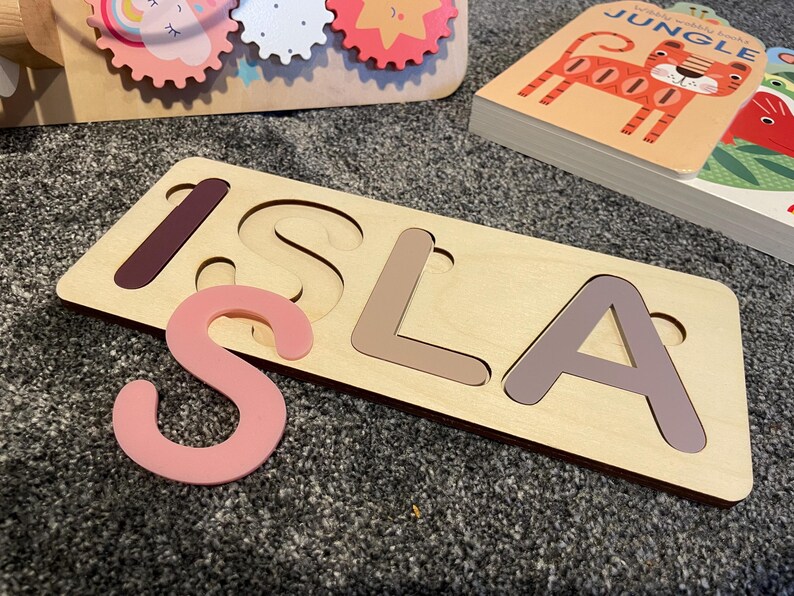 Puede incluir: Un rompecabezas de madera con el nombre "ISLA" recortado. Las letras son de diferentes tonos de marr&oacute;n y rosa. La "S" es una pieza separada y es rosa.