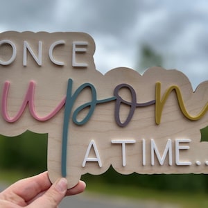 Könnte beinhalten: Holzschild mit dem Schriftzug "ONCE UPON A TIME..." in Weiß, Rosa, Türkis, Lila und Gelb. Das Schild ist in Form einer Wolke geschnitten. Der Hintergrund ist unscharf.