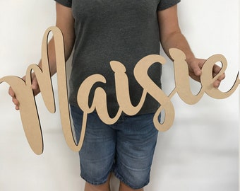 May include: A large, wooden sign with the name "Maisie" written in a cursive script. The sign is held by a person, and the wood is a light brown color.