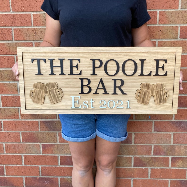 K&ouml;nnte beinhalten: Ein Holzschild mit dem Text "The Poole Bar Est 2021" in schwarzen Buchstaben. Das Schild hat zwei 3D-Bierkr&uuml;ge auf beiden Seiten des Textes.