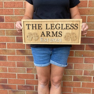 K&ouml;nnte beinhalten: Ein Holzschild mit dem Text "The Legless Arms Est 2021" und zwei geschnitzten Bierkr&uuml;gen.