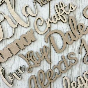 May include: A collection of wooden names, including "Love", "Alexia", "Dale", "Emma", "Crafts", and "Kayla", in a cursive font. The names are cut out of wood and have a natural brown colour.
