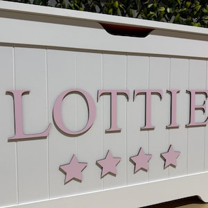 May include: White wooden toy chest featuring the name "LOTTIE" in pink lettering, with five pink star decorations. The chest is rectangular with a hinged lid. The background is a green hedge.