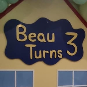 May include: A blue and yellow sign with the text "Beau Turns 3" in a wavy, cloud-like shape.