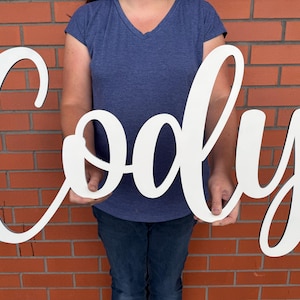 May include: A large, white wooden sign spelling out the name "Cody" in a cursive font. The sign is held in front of a brick wall. The letters have a black outline.