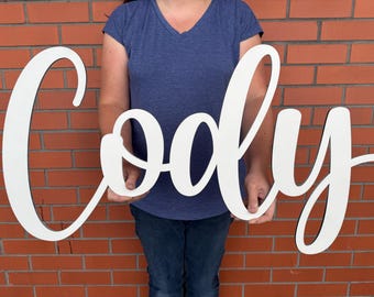 May include: A large, white wooden sign spelling out the name "Cody" in a cursive font. The sign is held in front of a brick wall. The letters have a black outline.