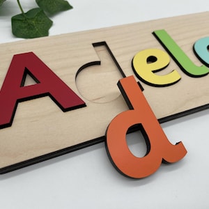 May include: A wooden name puzzle spelling "Adelaide" with colourful letters. The letters are red, yellow, green, and blue, with an orange "d" partially removed. The puzzle is made of light-coloured wood with a black border.