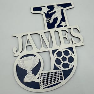 May include: A wooden sign with the name "JAMES" cut out in white. The design includes a soccer player kicking a ball, stars, a soccer ball, a trophy, and a goal, all with a navy blue background.