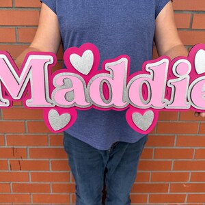 May include: A layered wooden sign spelling "Maddie" in pink and silver. The name is surrounded by pink hearts with silver glitter centers. The sign is held in front of a brick wall.