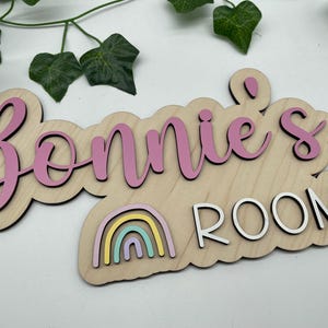 May include: Wooden sign with the name "Bonnie's" in pink script, and the word "ROOM" in white block letters. A colorful rainbow graphic is below the name. The sign is designed for a child's room.