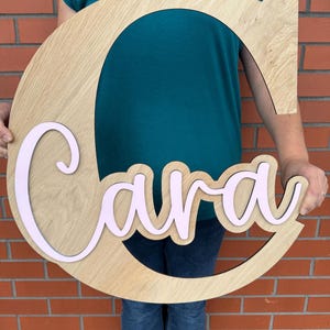 Peut inclure: Une lettre "C" en bois avec le nom "Cara" en rose clair. La lettre en bois est de couleur marron clair et présente un motif découpé. Le nom "Cara" est superposé à la lettre "C".
