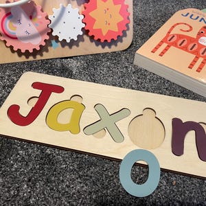 Puede incluir: Un rompecabezas de madera con el nombre Jaxon escrito en letras de colores. Las piezas del rompecabezas tienen la forma de las letras y encajan en los recortes correspondientes. Hay una letra O azul junto al rompecabezas.