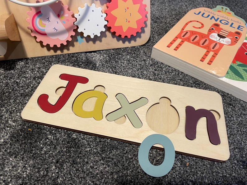Puede incluir: Un rompecabezas de madera con el nombre Jaxon escrito en letras de colores. Las piezas del rompecabezas tienen la forma de las letras y encajan en los recortes correspondientes. Hay una letra O azul junto al rompecabezas.