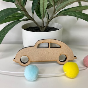 Könnte beinhalten: Ein hellbrauner Holzzuschnitt eines Autos mit kreisförmigen Rädern und Fensterdetails. Das Auto befindet sich vor einer Girlande aus pastellfarbenen Pompons. Ein weißer Topf mit einer grünen Pflanze befindet sich im Hintergrund.