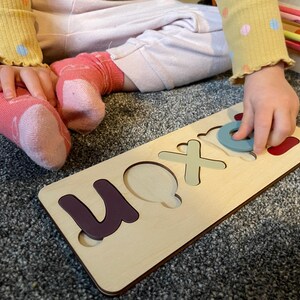 Puede incluir: Un tablero de rompecabezas de madera con recortes para las letras "U", "O", "X" y "N". Una mano de ni&ntilde;o est&aacute; colocando una pieza circular azul en el recorte "O".
