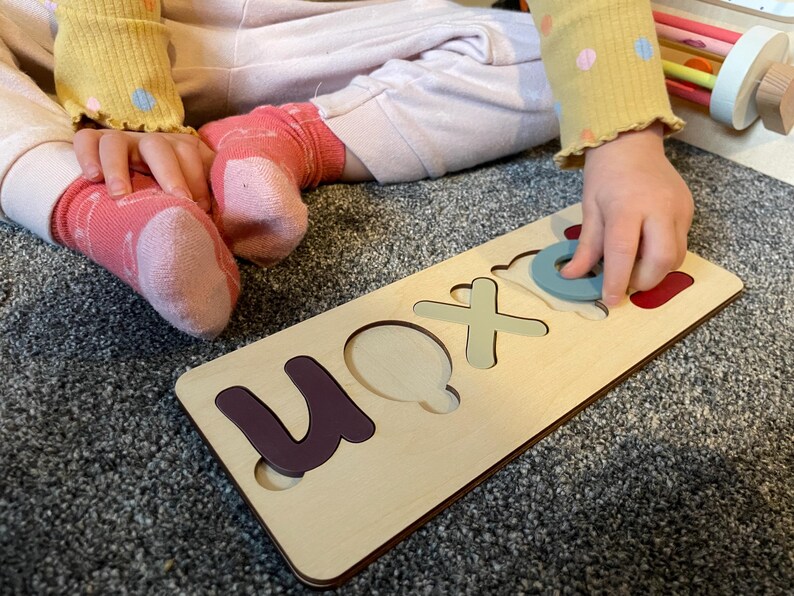 Puede incluir: Un tablero de rompecabezas de madera con recortes para las letras "U", "O", "X" y "N". Una mano de ni&ntilde;o est&aacute; colocando una pieza circular azul en el recorte "O".