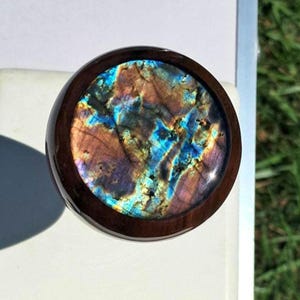 May include: A circular ring featuring a polished dark brown wooden band and a large, round, iridescent labradorite gemstone inlay. The stone displays vibrant flashes of electric blue, gold, and deep purple colors across its textured surface.
