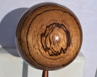 May include: A spherical wooden knob with a polished finish, showcasing prominent dark brown concentric grain patterns. The sphere is attached to a thin, vertical copper-colored metal rod extending downward from the center of the base.
