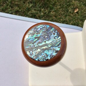 May include: Round wooden bottle stopper with a shimmering abalone shell inlay. The abalone shell features iridescent blues, greens, and purples. The stopper has a dark brown wooden rim and a smooth, polished finish.