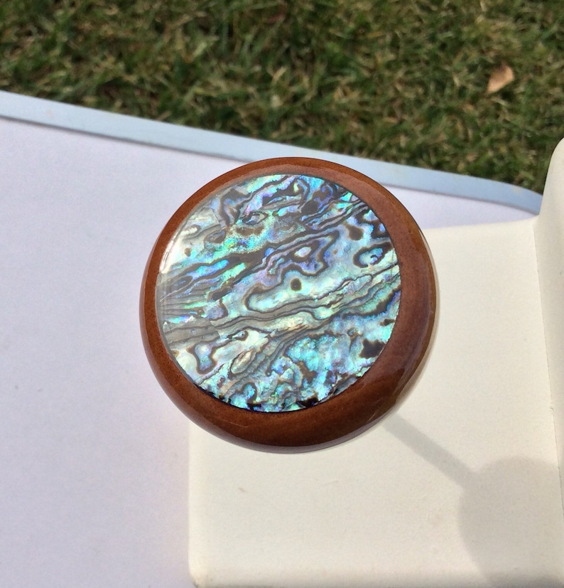May include: Round wooden bottle stopper with a shimmering abalone shell inlay. The abalone shell features iridescent blues, greens, and purples. The stopper has a dark brown wooden rim and a smooth, polished finish.