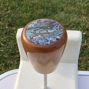May include: A wooden bottle stopper with a polished finish. The top features an abalone shell inlay with iridescent blues, greens, and purples. The stopper is mounted on a white stand, with a green grass background.