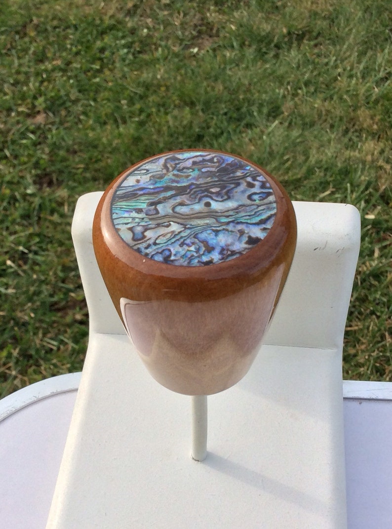 May include: A wooden bottle stopper with a polished finish. The top features an abalone shell inlay with iridescent blues, greens, and purples. The stopper is mounted on a white stand, with a green grass background.