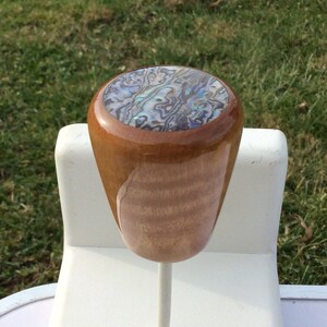 May include: A wooden shift knob with a polished finish. The top of the knob features an abalone inlay with iridescent blues, greens, and purples. The knob is mounted on a white metal rod, set against a backdrop of green grass.