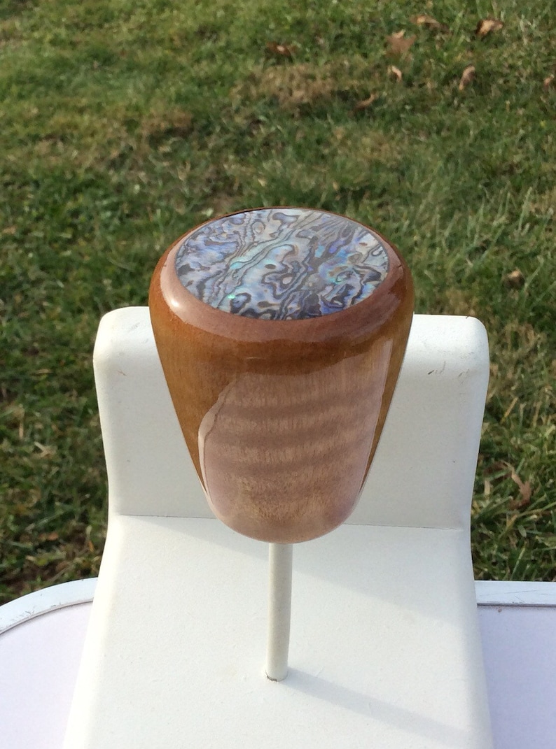 May include: A wooden shift knob with a polished finish. The top of the knob features an abalone inlay with iridescent blues, greens, and purples. The knob is mounted on a white metal rod, set against a backdrop of green grass.