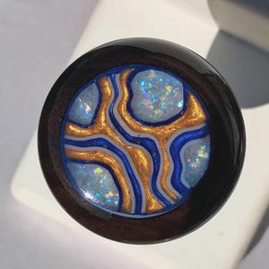 May include: A round, dark brown wooden plug with a central design. The design features a light blue background with gold and blue swirling patterns. The center has a glittery, iridescent effect.