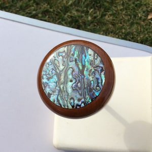 May include: A round wooden knob with a polished, dark brown finish. The center features a shimmering, iridescent abalone shell inlay with shades of blue, green, and silver. This decorative knob is suitable for furniture or cabinetry.